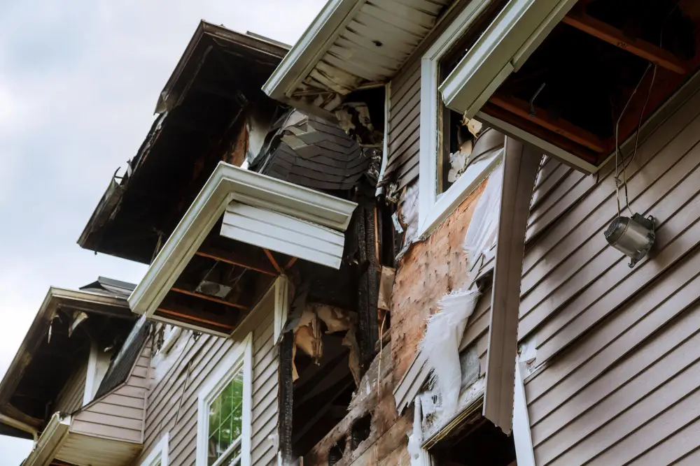 Severely damaged two story house with collapsed siding and exposed interior debris hanging from the roof edge